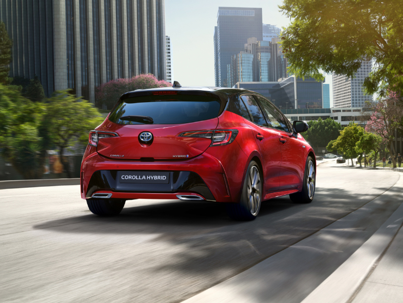 Zadní pohled na červený vůz Toyota Corolla Hybrid jedoucí po městské ulici.