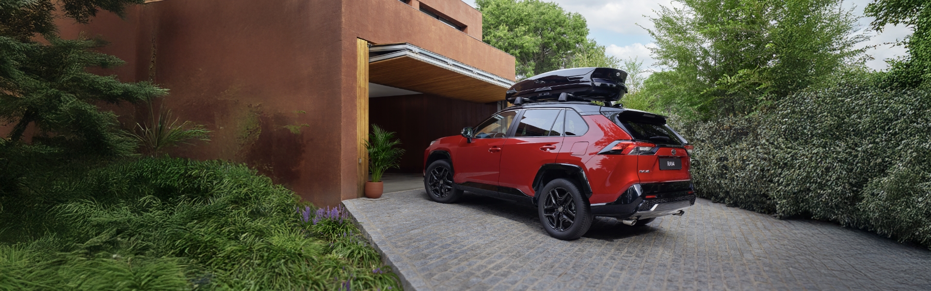 Červené SUV Toyota RAV4 s černým střešním boxem zaparkované na dlažbě před garáží.